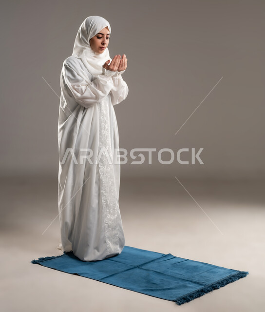ايماءات تدل على التوسل والتضرع، أداء الفروض على وقتها, التقرب الى الله من خلال الإجتهاد في العبادة والحفاظ على الصلوات، بورتريه لإمرأة عربية عمانية خليجية ترتدي الحجاب والعباءة البيضاء تقف على سجادة الصلاه ترفع يديها وتدعو الله, صورة الجسم بالطول الكامل, خلفية رمادية