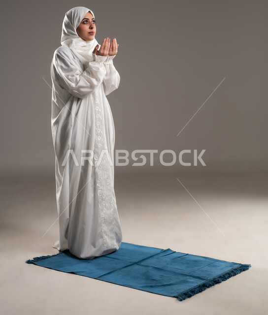 ايماءات تدل على التوسل والتضرع، أداء الفروض على وقتها, التقرب الى الله من خلال الإجتهاد في العبادة والحفاظ على الصلوات، بورتريه لإمرأة عربية عمانية خليجية ترتدي الحجاب والعباءة البيضاء تقف على سجادة الصلاه ترفع يديها وتدعو الله, صورة الجسم بالطول الكامل, خلفية رمادية