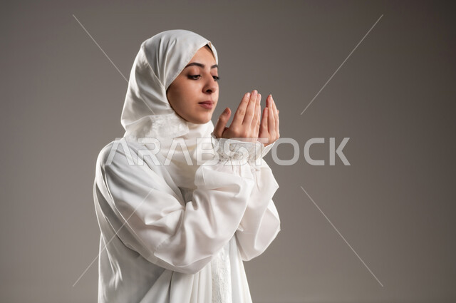 Gestures that indicate pleading and pleading, performing the obligations on time, getting closer to God through diligence in worship and observing prayers, a close-up portrait of an Omani Gulf Arab woman wearing the hijab and white abaya, standing on the prayer rug, raising her hands and praying to God, gray background.