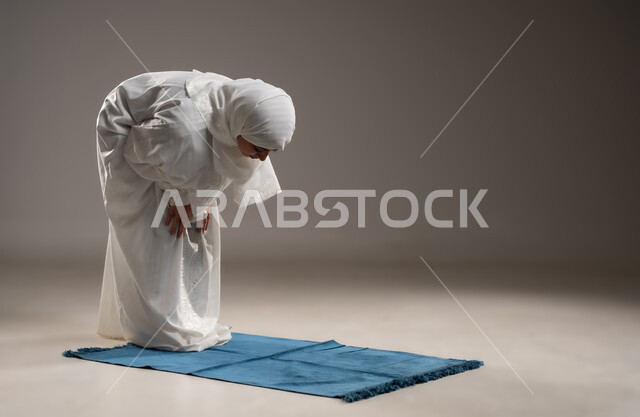 ايماءات تدل على الخشوع في الصلاة, أداء الفروض على وقتها, التقرب الى الله من خلال الإجتهاد في العبادة والحفاظ على الصلوات، بورتريه لإمرأة عربية عمانية خليجية ترتدي الحجاب والعباءة البيضاء تركع على سجادة الصلاة,  خلفية رمادية