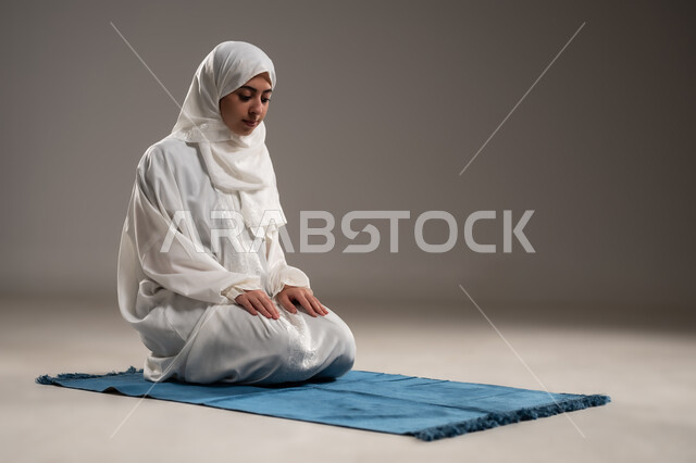 ايماءات تدل على الخشوع في الصلاة, أداء الفروض على وقتها, التقرب الى الله من خلال الإجتهاد في العبادة والحفاظ على الصلوات، بورتريه  لإمرأة عربية عمانية خليجية ترتدي الحجاب والعباءة البيضاء تلجس على سجادة الصلاة, صورة الجسم بالطول الكامل, خلفية رمادية