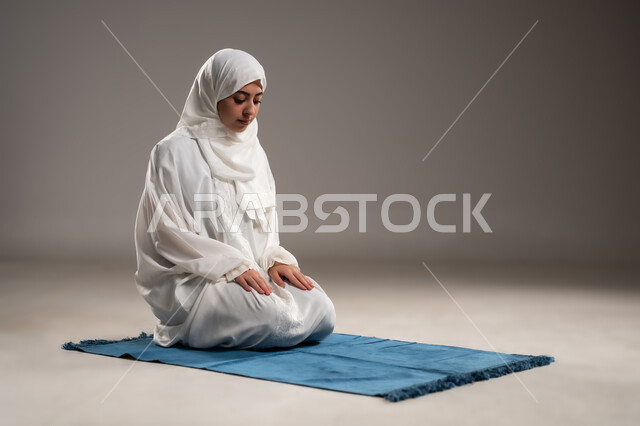 ايماءات تدل على الخشوع في الصلاة, أداء الفروض على وقتها, التقرب الى الله من خلال الإجتهاد في العبادة والحفاظ على الصلوات، بورتريه  لإمرأة عربية عمانية خليجية ترتدي الحجاب والعباءة البيضاء تلجس على سجادة الصلاة, صورة الجسم بالطول الكامل, خلفية رمادية