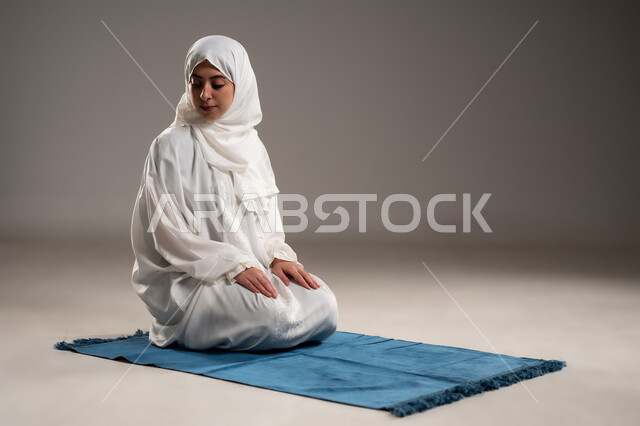 ايماءات تدل على الخشوع في الصلاة, أداء الفروض على وقتها, التقرب الى الله من خلال الإجتهاد في العبادة والحفاظ على الصلوات، بورتريه  لإمرأة عربية عمانية خليجية ترتدي الحجاب والعباءة البيضاء تلجس على سجادة الصلاة, صورة الجسم بالطول الكامل, خلفية رمادية