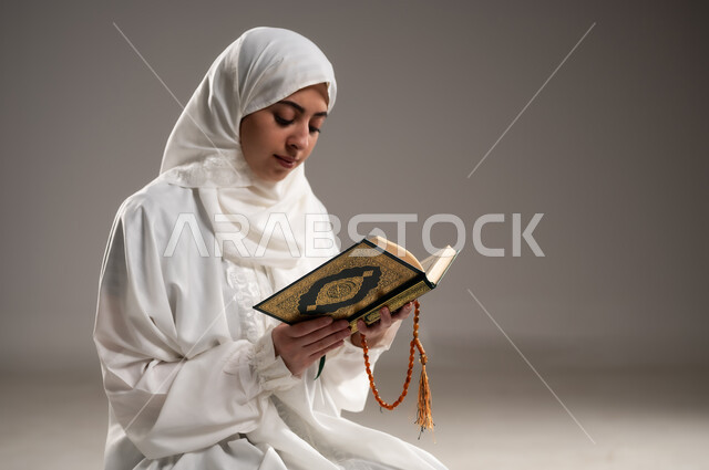 الصلاه وقراءة القرآن, بورتريه مقرب لإمرأة عربية عمانية خليجية ترتدي الحجاب والعباءة البيضاء تجلس على سجادة الصلاه تقرأ القرآن, ذكر الله تعالى و الدعاء والإستغفار, الإجتهاد في العباده في شهر رمضان المبارك, التقرب الى الله تعالى, خلفية رمادية