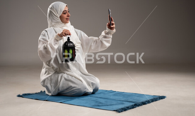 مفهوم الاحتفال بقدوم المناسبات الدينية، التقاط الصور التذكارية, فوانيس اسلامية للاعياد في سلطنه عمان, الالتزام بأداء الفروض على وقتها, التقرب الى الله بالإجتهاد في العبادة والحفاظ على الصلوات، بورتريه لإمرأة عربية عمانية خليجية ترتدي الحجاب والعباءة البيضاء, الجلوس على سجادة الصلاة وحمل فانوس رمضاني وهاتف محمول، خلفية رمادية