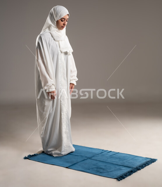 ايماءات تدل على الخشوع في الصلاة, أداء الفروض على وقتها,  التقرب الى الله من خلال الإجتهاد في العبادة والحفاظ على الصلوات، بورتريه من الخلف لإمرأة عربية عمانية خليجية ترتدي الحجاب والعباءة البيضاء تقف على سجادة الصلاة, صورة الجسم بالطول الكامل, خلفية رمادية
