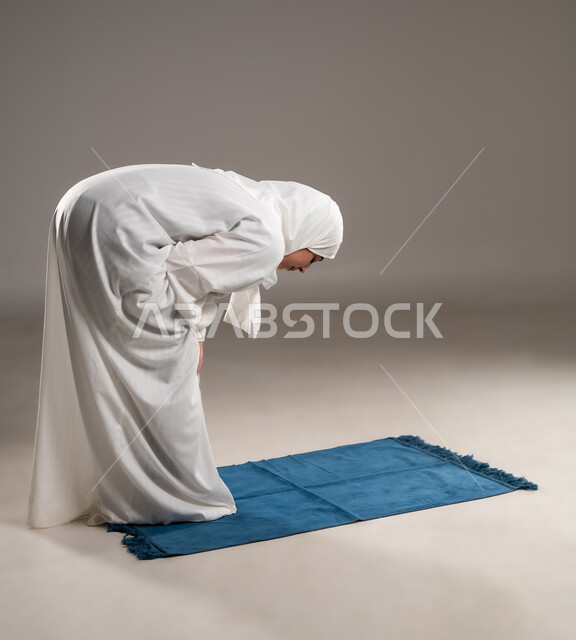 أداء الفروض على وقتها, ايماءات تدل على الخشوع في الصلاة, التقرب الى الله من خلال الإجتهاد في العبادة والحفاظ على الصلوات، بورتريه من الخلف لإمرأة عربية عمانية خليجية ترتدي الحجاب والعباءة البيضاء تركع على سجادة الصلاة, خلفية رمادية