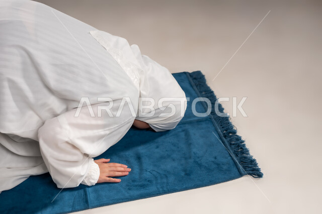 أداء الفروض على وقتها, ايماءات تدل على الخشوع في الصلاة, التقرب الى الله من خلال الإجتهاد في العبادة والحفاظ على الصلوات، بورتريه مقرب لإمرأة عربية عمانية خليجية ترتدي الحجاب والعباءة البيضاء تسجد على سجادة الصلاة, خلفية رمادية