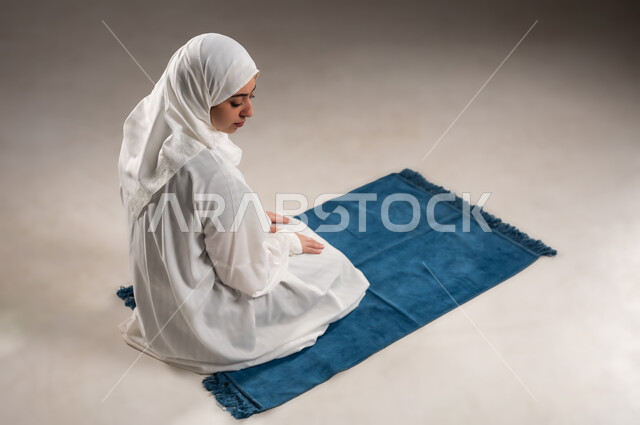 ايماءات تدل على الخشوع في الصلاة, أداء الفروض على وقتها, التقرب الى الله من خلال الإجتهاد في العبادة والحفاظ على الصلوات، بورتريه لإمرأة عربية عمانية خليجية ترتدي الحجاب والعباءة البيضاء تجلس على السجادة في وقت الصلاة, خلفية رمادية