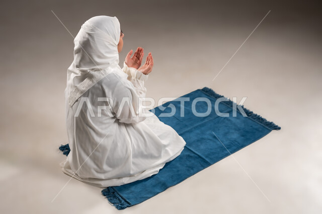 ايماءات تدل على التوسل والتضرع، أداء الفروض على وقتها, التقرب الى الله من خلال الإجتهاد في العبادة والحفاظ على الصلوات، بورتريه  لإمرأة عربية عمانية خليجية ترتدي الحجاب والعباءة البيضاء تجلس على سجادة الصلاه ترفع يديها وتدعو الله, خلفية رمادية