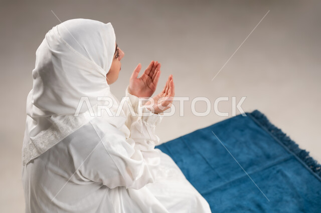Gestures indicating pleading and supplication, performing the obligations on time, getting closer to God through diligence in worship and observing prayers, a close-up portrait of an Omani Gulf Arab woman wearing the hijab and white abaya, sitting on the prayer rug, raising her hands and praying to God, gray background.