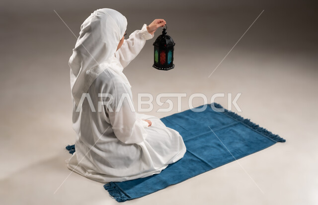 مفهوم الاحتفال بقدوم المناسبات الدينية، فوانيس اسلامية للاعياد في سلطنه عمان, الالتزام بأداء الفروض على وقتها, التقرب الى الله بالإجتهاد في العبادة والحفاظ على الصلوات، بورتريه لإمرأة عربية عمانية خليجية ترتدي الحجاب والعباءة البيضاء, الجلوس على سجادة الصلاة وحمل فانوس رمضاني، خلفية رمادية