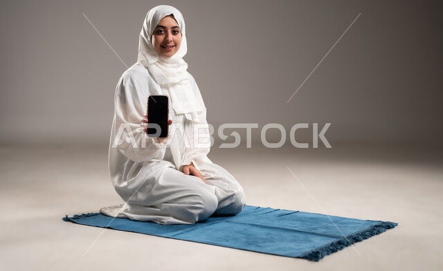 The concept of the pillars of Islam and faith, commitment to performing obligations on time, getting closer to God through diligence in worship and observing prayers, portrait of an Omani Gulf Arab woman wearing the hijab and white abaya, sitting on a prayer rug and showing a mobile phone with a black screen, gray background