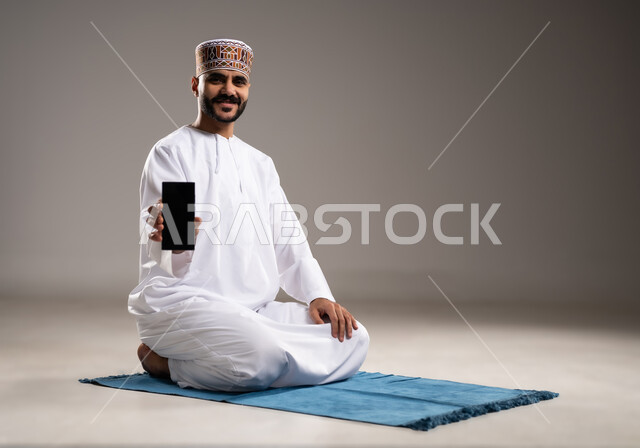 التقرب من الله تعالى بأداء الفرائض والتزام أوامره, الركن الثاني من اركان الاسلام، بورتريه مقرب لرجل عربي خليجي عماني يرتدي الدشداشة والكمة, الجلوس على سجادة الصلاة وعرض هاتف محمول بشاشة سوداء، الحفاظ على إقامة الصلوات الخمس، خلفية رمادية