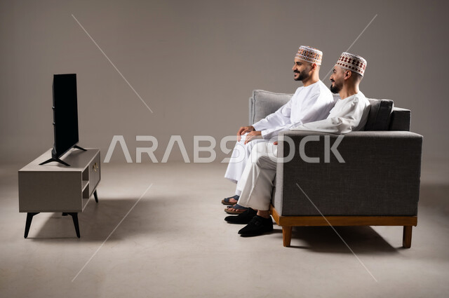 المباريات والالعاب الرياضية, التفاعل والاندماج بمشاهدة الفريق المفضل, ايماءات الفرح والحماس بالفوز والانتصار, بورتريه لرجلان عربيان عمانين خليجيان يرتديان الدشداشة والكمة ويجلسون على الاريكه, الشغف بالانشطة الترفيهية, خلفية رمادية