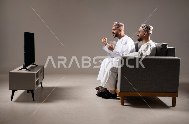 المباريات والالعاب الرياضية, التفاعل والاندماج بمشاهدة الفريق المفضل, ايماءات الفرح والحماس بالفوز والانتصار, بورتريه لرجلان عربيان عمانين خليجيان يرتديان الدشداشة والكمة ويجلسون على الاريكه, الشغف بالانشطة الترفيهية, خلفية رمادية