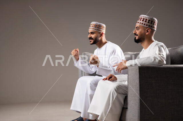 المباريات والالعاب الرياضية, التفاعل والاندماج بمشاهدة الفريق المفضل, ايماءات الفرح والحماس بالفوز والانتصار, بورتريه لرجلان عربيان عمانين خليجيان يرتديان الدشداشة والكمة ويجلسون على الاريكه, الشغف بالانشطة الترفيهية, خلفية رمادية