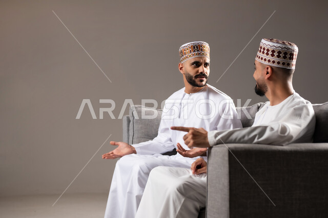 الاستمتاع بتبادل الاحاديث مع الاصدقاء, مفهوم العلاقة القوية والترابط بين الزملاء, تعابير التسلية والمرح, بورتريه لرجلان عربيان عمانيان خليجيان يرتدون الدشداشة مع الكمه ويجلسون على الاريكه, نشاط منزلي ترفيهي جماعي, قضاء اوقات ممتعة ومسلية, خلفية رمادية