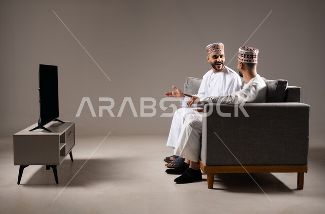 الاستمتاع بتبادل الاحاديث مع الاصدقاء, مفهوم العلاقة القوية والترابط بين الزملاء, تعابير التسلية والمرح, بورتريه لرجلان عربيان عمانيان خليجيان يرتدون الدشداشة مع الكمه ويجلسون على الاريكه, نشاط منزلي ترفيهي جماعي, قضاء اوقات ممتعة ومسلية, خلفية رمادية
