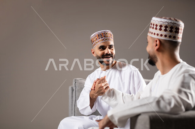 الاستمتاع بتبادل الاحاديث مع الاصدقاء, مفهوم العلاقة القوية والترابط بين الزملاء, تعابير التسلية والمرح, بورتريه لرجلان عربيان عمانيان خليجيان يرتدون الدشداشة مع الكمه ويجلسون على الاريكه, نشاط منزلي ترفيهي جماعي, قضاء اوقات ممتعة ومسلية, خلفية رمادية
