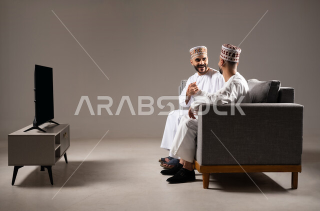 الاستمتاع بتبادل الاحاديث مع الاصدقاء, مفهوم العلاقة القوية والترابط بين الزملاء, تعابير التسلية والمرح, بورتريه لرجلان عربيان عمانيان خليجيان يرتدون الدشداشة مع الكمه ويجلسون على الاريكه, نشاط منزلي ترفيهي جماعي, قضاء اوقات ممتعة ومسلية, خلفية رمادية