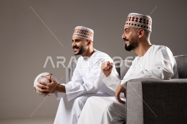 التفاعل والاندماج بمشاهدة الفريق المفضل, المباريات والالعاب الرياضية, ايماءات الفرح والحماس بالفوز والانتصار, بورتريه لرجلان عربيان عمانين خليجيان يرتديان الدشداشة والكمة احدهما يمسك بيده كرة قدم, الشغف بالانشطة الترفيهية, خلفية رمادية