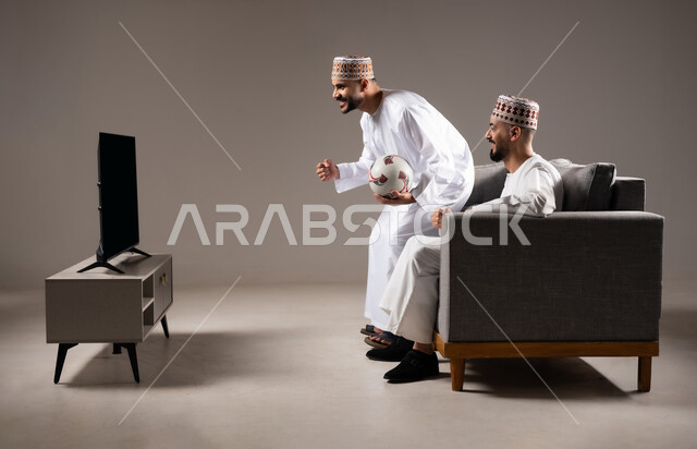 التفاعل والاندماج بمشاهدة الفريق المفضل, المباريات والالعاب الرياضية, ايماءات الفرح والحماس بالفوز والانتصار, بورتريه لرجلان عربيان عمانين خليجيان يرتديان الدشداشة والكمة احدهما يمسك بيده كرة قدم, الشغف بالانشطة الترفيهية, خلفية رمادية