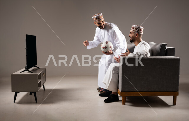 التفاعل والاندماج بمشاهدة الفريق المفضل, المباريات والالعاب الرياضية, ايماءات الفرح والحماس بالفوز والانتصار, بورتريه لرجلان عربيان عمانين خليجيان يرتديان الدشداشة والكمة احدهما يمسك بيده كرة قدم, الشغف بالانشطة الترفيهية, خلفية رمادية