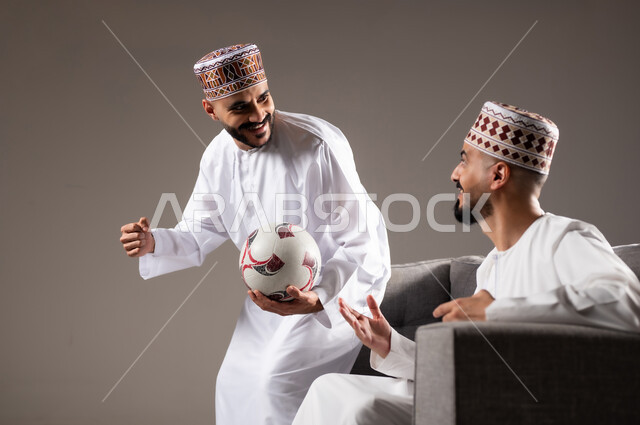 Interaction and integration by watching the favorite team, sports matches and games, gestures of joy and enthusiasm for victory and triumph, portrait of two Omani Gulf Arab men wearing a dishdasha and a kabbah, one of them holding a football in his hand, passion for recreational activities, gray background