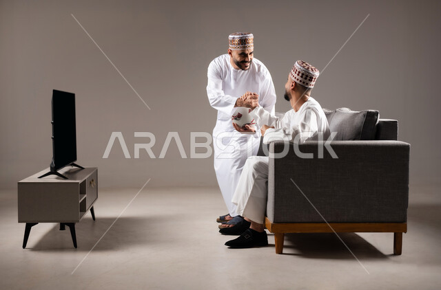 Interaction and integration by watching the favorite team, sports matches and games, gestures of joy and enthusiasm for victory and triumph, portrait of two Omani Gulf Arab men wearing a dishdasha and a kabbah, one of them holding a football in his hand, passion for recreational activities, gray background