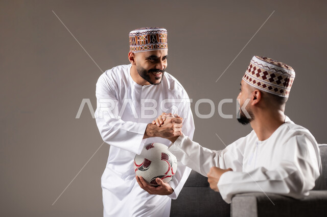 Interaction and integration by watching the favorite team, sports matches and games, gestures of joy and enthusiasm for victory and triumph, portrait of two Omani Gulf Arab men wearing a dishdasha and a kabbah, one of them holding a football in his hand, passion for recreational activities, gray background