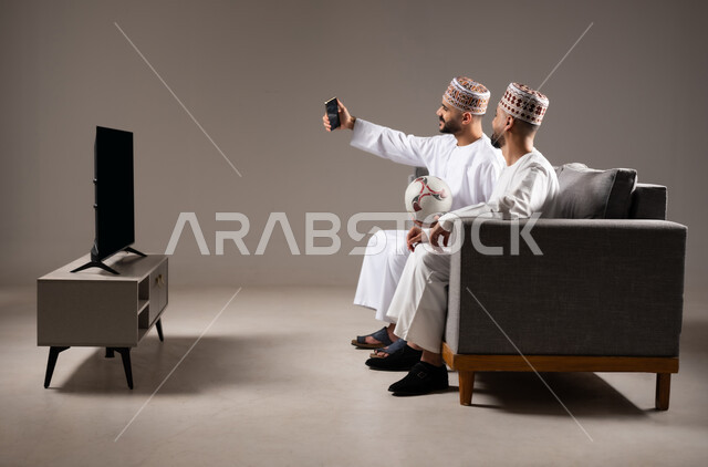 التقاط الصور التذكارية,  قضاء وقت ممتع مع العائلة والاصدقاء, ايماءات الفرح والسرور والسعادة, بورتريه لرجلان عربيان عمانين خليجيان يرتديان الدشداشة والكمة يمسك احدهم هاتف محمول ويلتقط صورة, خلفية رمادية