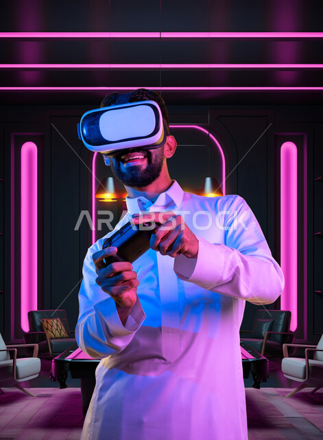 محاكاة العالم الخيالي، تعابير الاندماج والتفاعل، بورتريه مقرب لشاب عربي سعودي خليجي يرتدي الثوب التقليدي ونظارات الواقع الافتراضي يحمل عصا التحكم يقوم باللعب في ألعاب الفيديو ينظر الى الكاميرا بإيماءات الحماس، اجهزة وتقنيات حديثة, خلفية ملونة