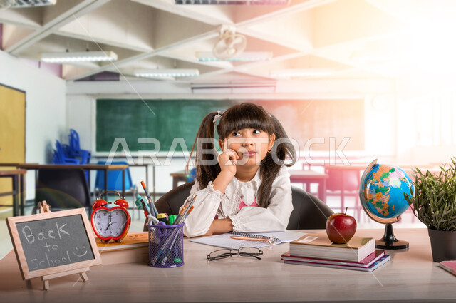 Preparing for the new school year, back to school, a Saudi Arabian Gulf student wearing a school uniform sitting inside the classroom looking at something with gestures of thinking and concentration, the concept of diligence and preparing for exams, education in schools in the Kingdom of Saudi Arabia