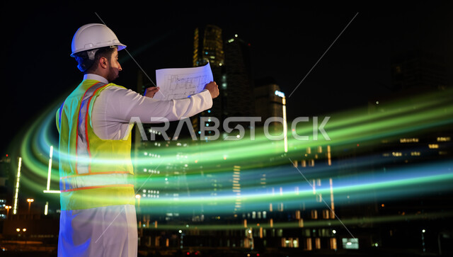 متابعة الأعمال الهندسية, المخططات الانشائية المعمارية, تنمية وتطور السعودية على يد أبناء الوطن, صورة من الخلف لمهندس عربي خليجي سعودي يرتدي الخوذة وسترة الحماية يمسك بيده المخطط يقف في موقع العمل ليلا، الأضواء والخطوط الهولوجرامية