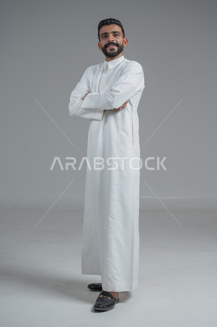 الاهتمام بالمظهر الخارجي والأناقة، مفهوم الرجولة والثقة بالنفس، بورتريه لرجل عربي خليجي سعودي يرتدي الثوب التقليدي ينظر الى الكاميرا بتعابير السرور، الوقوف باستقامة وثبات بايدي متقاطعة، صورة الجسم بالطول الكامل، خلفية رمادية