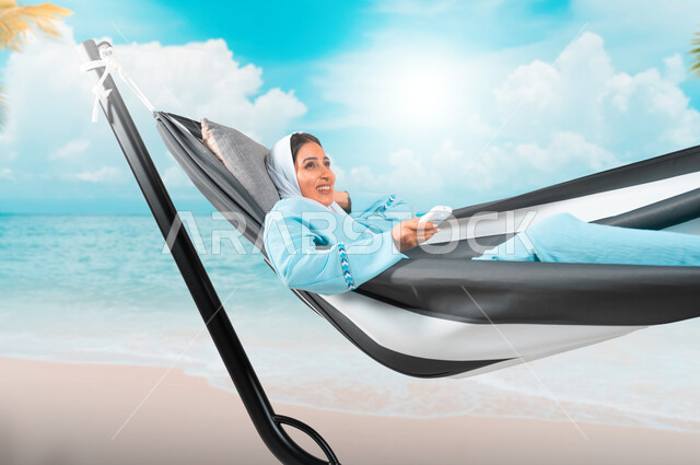 قضاء اوقات ممتعة في الاستراحة، الشعور بالاسترخاء والراحة والسكون، شابة عربية خليجية سعودية ترتدي لباس الكاجوال الخاص بالرحلات الصيفية مستلقية على أرجوحة قماشية تمسك بيدها جهاز تحكم، التخطيط للعطلة الصيفية بإيماءات السرور