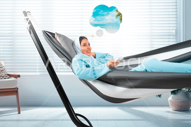 قضاء اوقات ممتعة في الاستراحة، الشعور بالاسترخاء والراحة والسكون، شابة عربية خليجية سعودية ترتدي لباس الكاجوال الخاص بالرحلات الصيفية مستلقية على أرجوحة قماشية تمسك بيدها جهاز تحكم، التخطيط للعطلة الصيفية بإيماءات السرور