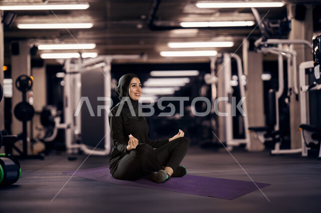 الشعور بالراحة و الاسترخاء، الاستمتاع في قضاء الوقت بالتأمل، متابعة تمارين اليوغا في النادي الرياضي، المحافظة على اللياقة والصحة البدنية، شابة عربية خليجية سعودية محجبة ترتدي الزي الرياضي تقوم بممارسة اليوغا بإيماءات السرور، التدرب على الهدوء وتمالك النفس