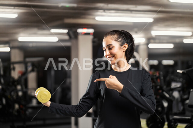 قضاء الأوقات في ممارسة الرياضة, صورة مقربة لامرأة عربية خليجية سعودية ترتدي ملابس رياضية تشير بإصبع السبابة الى يدها الثانية تقوم برفع الأوزان بإيماءات السرور, اتباع اسلوب حياة صحي, مفهوم الرشاقة واللياقة، تمارين تقوية العضلات حرق الدهون