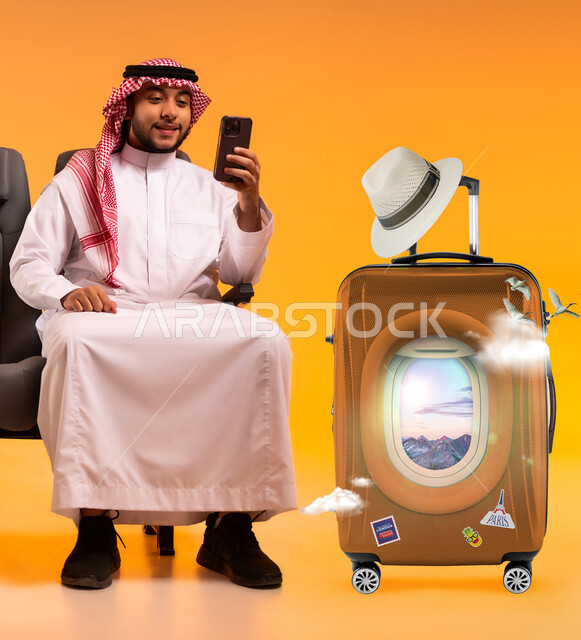 الحجوزات الالكترونية للرحلات السياحية, الاستعداد لقضاء وقت ممتع في العطلة، بورتريه لشاب عربي خليجي سعودي مبتسم يرتدي الشماغ و الثوب التقليدي يجلس على الكرسي بجانب حقيبة الامتعة يحمل بيده الهاتف المحمول, تعابير الفرح والسرور والحماس، خلفية صفراء