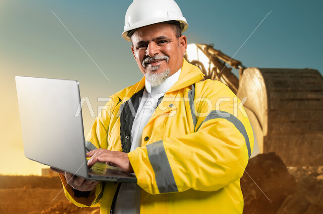 دمج التكنولوجيا الحديثة بالأعمال الهندسية, استخدام جهاز الحاسوب لإنجاز المهام, مفهوم الهندسة المعمارية, صورة مقربة لمهندس كبير السن عربي خليجي سعودي يرتدي الخوذة و سترة الحماية يمسك بيده الحاسب المحمول ينظر الى الكاميرا بإيماءات السرور, الإشراف على سير العمل