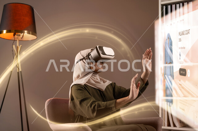 محاكاة العالم الخيالي، تعابير الاندماج والتفاعل مع الواقع الافتراضي، صورة مقربة لسيدة عربية خليجية سعودية محجبة تجلس على كرسي مريح ترتدي نظارات الفي آر تحاول لمس العناصر الهولوجرامية المضيئة، مفهوم التطور الرقمي والتكنولوجي