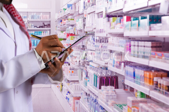 فحص وجرد مخزون الأدوية والمستلزمات الطبية داخل الصيدلية، تقديم الرعاية الصحية والوصفات الطبية، صورة مقربة ليد صيدلاني عربي سعودي خليجي يرتدي اليونيفورم الطبي يقوم بمراجعة الفواتير وتسجيل النقص في الدواء، مهن ووظائف سعودية