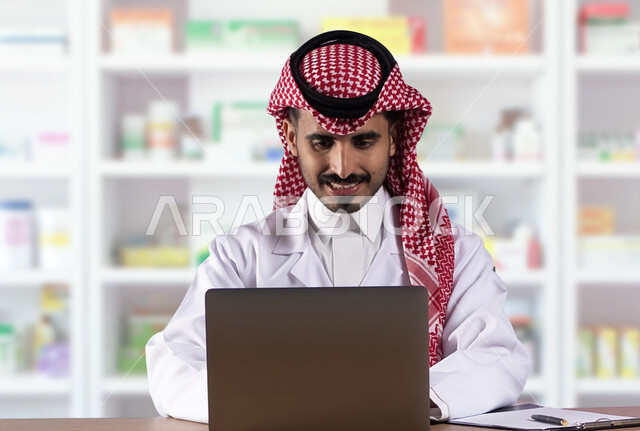 استخدام التقنيات والأجهزة الحديثة، فحص وجرد مخزون الأدوية والمستلزمات الطبية داخل الصيدلية، صورة مقربة لصيدلاني عربي سعودي خليجي يرتدي الشماغ والثوب التقليدي يعمل على جهاز اللابتوب المحمول بإيماءات السرور، مهن ووظائف سعودية