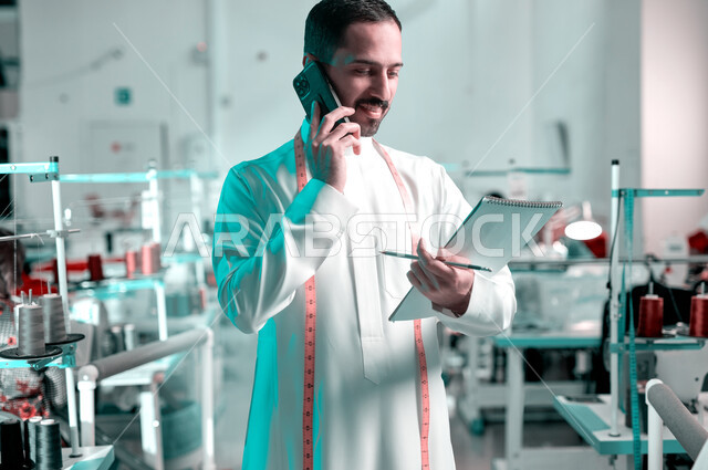 إتقان واحتراف صنع الملابس، تسجيل وتدوين القياسات على الدفتر، صورة مقربة لخياط عربي خليجي سعودي يرتدي الثوب التقليدي يضع على رقبته الشريط المتري يقوم بإجراء مكالمة هاتفية، استعمال أدوات ومعدات الخياطة، استخدام التقنيات والأجهزة الحديثة