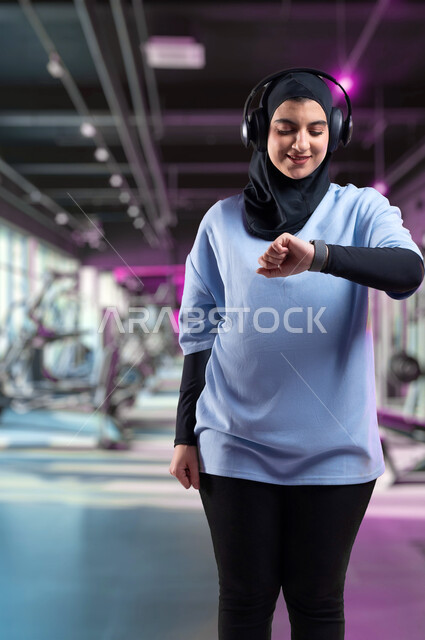 التدرب بعزيمة للمحافظة على نظام الحياة الصحي، ايماءات السعادة والنشاط، مفهوم الرشاقة واللياقة، بورتريه لفتاة عربية خليجية سعودية محجبة ترتدي الملابس الرياضية تضع الهيدفون تتابع تقدمها في الأداء على ساعة اليد، مراقبة الوقت لبدء التمرين، ممارسة الهوايات المفيدة