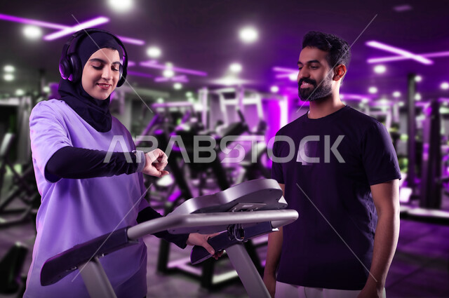 مفهوم حرق الدهون و إنقاص الوزن، أداء التمرينات بشكل منتظم، انشطة رياضية لتقوية العضلات ونحت الجسم، مدرب عربي خليجي سعودي يرتدي الملابس الرياضية يقوم بتدريب امرأة على جهاز العجلة الهوائية, شابة تقوم بمتابعة تقدمها على ساعة اليد الالكترونية