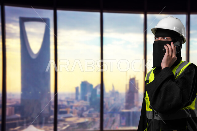 نهضة المجتمع السعودي بسواعد أبنائه، وظائف نسائية هندسية في المملكة, دمج التقنيات التكنولوجية بالمجال الهندسي، صورة مقربة لمهندسة عربية خليجية سعودية منقبة ترتدي الخوذة البيضاء وسترة الحماية تمسك الهاتف المحمول تقوم بإجراء مكالمة هاتفية، خلفية برج المملكة