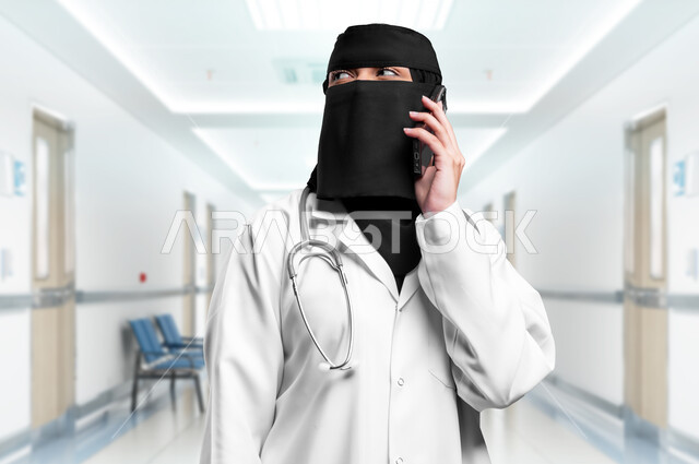 العمل في القطاع الصحي, استخدام التقنيات والأجهزة الحديثة، صورة مقربة لطبيبة عربية خليجية سعودية محجبة ترتدي المعطف الطبي الأبيض والنقاب تمسك بيدها الهاتف المحمول تقوم بإجراء مكالمة صوتية، تقديم الخدمات العلاجية والفحوصات الطبية للمرضى